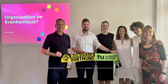 Group photo of six people in a seminar room. Two people are holding a scarf together that features the logos of Borussia Dortmund and TU Dortmund. In the background on the left, a presentation slide can be seen with the heading “Organization in a Hospital?” and the name “Marc Detzler.”