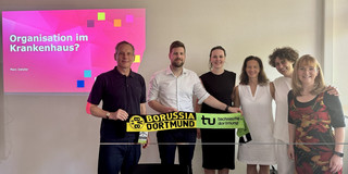 Group photo of six people in a seminar room. Two people are holding a scarf together that features the logos of Borussia Dortmund and TU Dortmund. In the background on the left, a presentation slide can be seen with the heading “Organization in a Hospital?” and the name “Marc Detzler.”