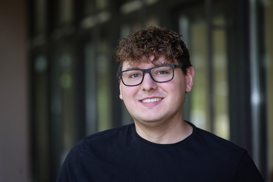 A portrait photo of Dustin Cirkel with short, curly hair and dark glasses. He is wearing a black top and looking at the camera. In the background, the blurred outlines of windows and walls in an interior space are visible.