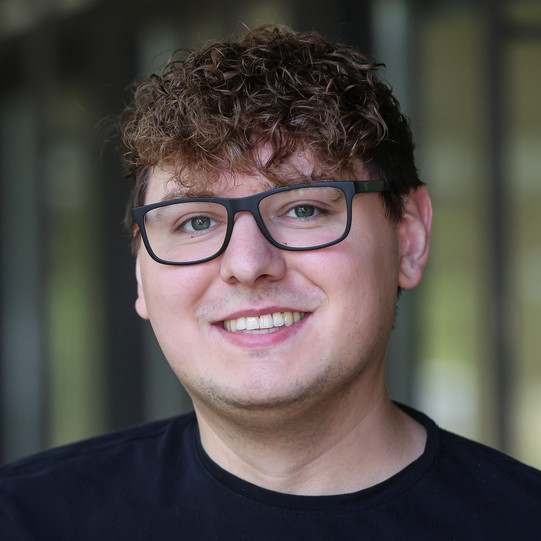 A portrait photo of Dustin Cirkel with short, curly hair and dark glasses. He is wearing a black top and looking at the camera. In the background, the blurred outlines of windows and walls in an interior space are visible.