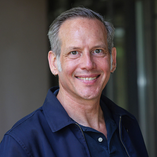 A portrait photo of Prof. Dr. Cornelius Schubert taken outdoors. He is wearing a dark blue polo shirt and a dark blue vest. Blurred windows and walls can be seen in the background.