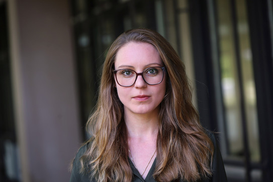 Porträtfoto von Elena Rosenberg mit langen, hellbraunen Haaren und dunkler Brille. Sie trägt ein dunkles Oberteil und blickt direkt in die Kamera. Im Hintergrund sind unscharfe Fenster- und Wandflächen eines Innenraums zu sehen.