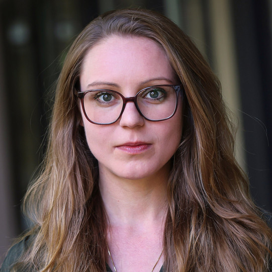 A portrait photo of Elena Rosenberg with long, light brown hair and dark glasses. She is wearing a dark top and looking directly at the camera. In the background, the blurred outlines of windows and walls in an interior space are visible.
