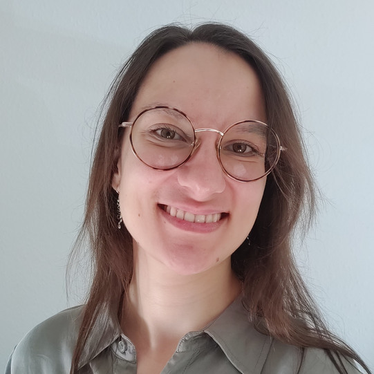 Porträt von Joana Rutz mit langen braunen Haaren und runder goldener Brille, lächelnd, in einem hellgrauen Hemd vor hellem, einfarbigem Hintergrund.