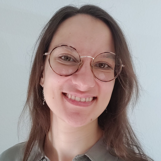 A portrait of Joana Rutz with long brown hair and round gold-rimmed glasses, smiling, wearing a light gray shirt against a light, solid-color background.