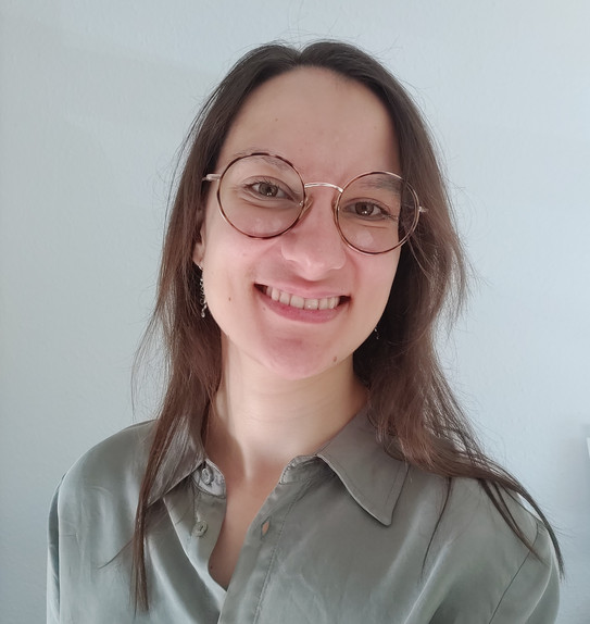 A portrait of Joana Rutz with long brown hair and round gold-rimmed glasses, smiling, wearing a light gray shirt against a light, solid-color background.