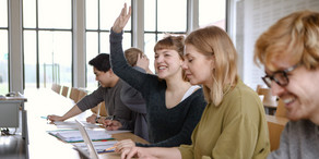 Fünf Studierende sitzen in einem Seminarraum an Tischen. Eine Studentin hebt ihre Hand.